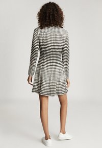Schwarz-weiß gemustertes Kleid mit langen Ärmeln, ausgestelltem Rock und strukturiertem Stoff. Weiße Sneakers ergänzen das Outfit.