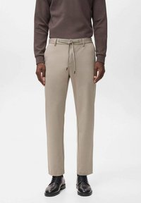 Beige Hosen mit geradem Schnitt, elastischem Bund und Kordelzug. Das Material wirkt glatt mit subtiler Textur. Kombiniert mit schwarzen Schuhen.