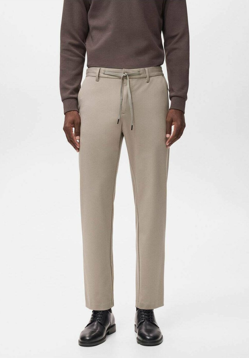 Beige broek met een rechte pasvorm, elastische tailleband en koord. Het materiaal lijkt glad met een subtiele textuur. Gecombineerd met zwarte schoenen.