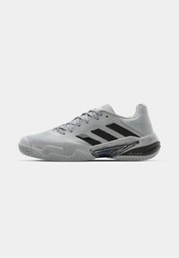 Hellgrauer Sportschuh mit schwarzen Streifen, ausgestattet mit einem strukturierten Obermaterial, gepolstertem Kragen und einem transparenten Akzent an der Seite.