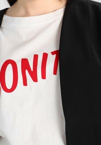 Musta bleiseri valkoisen T-paidan päällä, jossa on rohkeilla punaisilla kirjaimilla teksti "DONIT". Kangas näyttää sileältä ja siinä on jämäkkä istuvuus.
