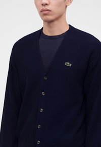 Homme portant un cardigan bleu marine foncé boutonné sur une chemise marine assortie avec un logo crocodile vert sur la poitrine gauche.