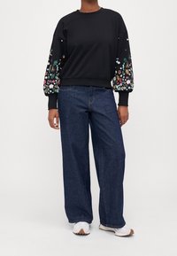 Sweat-shirt noir avec des motifs floraux brodés sur les manches bouffantes, associé à un jean en denim foncé ample et des baskets blanches.