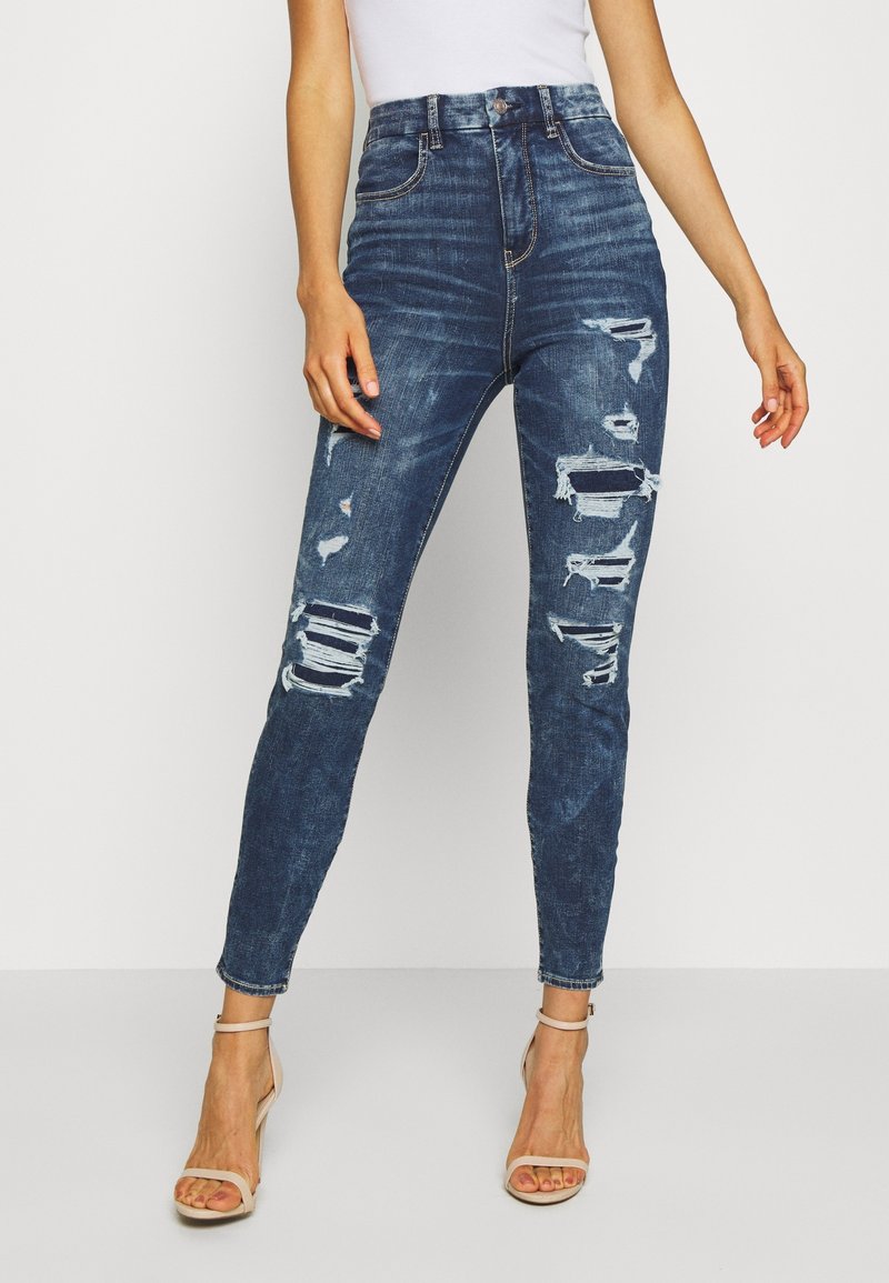 Jeans ajustados de tiro alto en denim azul oscuro. Presentan detalles desgastados con desgarros y bordes deshilachados. Corte ajustado, de longitud hasta el tobillo.