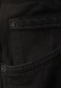 Gros plan sur un tissu en denim noir montrant des coutures détaillées et des rivets sur une poche avant.