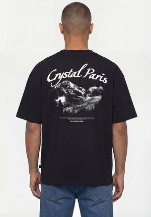 Mann trägt ein schwarzes T-Shirt mit dem Text „Crystal Paris“ und Berggrafik auf dem Rücken, kombiniert mit blauen Jeans, steht mit dem Rücken zum Betrachter.