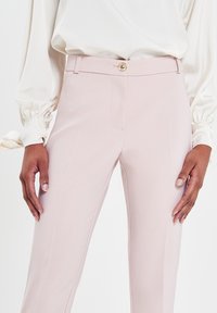 Helle rosa maßgeschneiderte Hose aus glattem Stoff, mit einem Knopfverschluss vorne und seitlichen Taschen. Der Bund ist schmal und elegant.