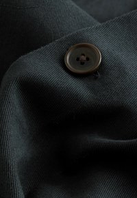 Tmavý textil s textúrovaným povrchom, na ktorom sa nachádza hnedý gombík so štyrmi dierkami. Materiál vyzerá hladko a je mierne ryhovaný.