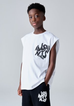 Chlapec v bielom tielku a čiernych šortkách s logom "AllSaints London" pózuje na obyčajnom sivom pozadí.