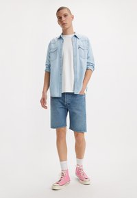 Light blue denim shirt over a white t-shirt, paired with blue denim shorts and pink high-top sneakers. Features rolled-up sleeves and pockets.