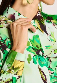 Vrouw die een knoop dichtdoet op een groene blouse met bloemenprint en lange, manchetachtige mouwen, met zichtbare gouden hoopsieraden.