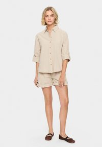 Beige linen shirt with rolled sleeves, button front; paired with matching beige shorts. Worn with brown flat shoes, set against a light background.