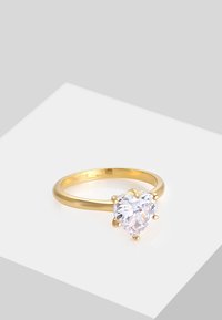 Anello placcato in oro con pietra preziosa trasparente a forma di cuore, fissata da quattro griffe su una fascia liscia. Marcatura della taglia visibile all'interno.