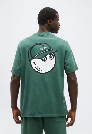 Homme portant une chemise verte avec un graphisme d'une balle de golf portant une casquette verte avec le texte "oralbon" au dos, vu de profil.