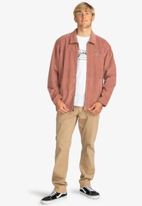 Giacca in corduroy di colore rosa polveroso, con colletto, chiusura con zip e tasche laterali, indossata sopra una maglietta bianca con grafica e pantaloni beige.