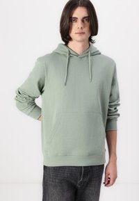 Felpa con cappuccio verde chiaro in tessuto morbido. Presenta una tasca frontale, polsini a costine e una vestibilità rilassata. Abbinata a jeans scuri.