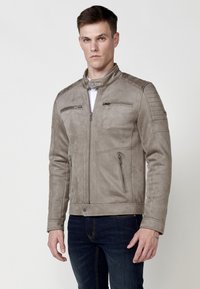 Jachetă din suede gri, cu un design ajustat, care prezintă două buzunare cu fermoar pe piept, buzunare laterale și accente matlasate pe umeri.
