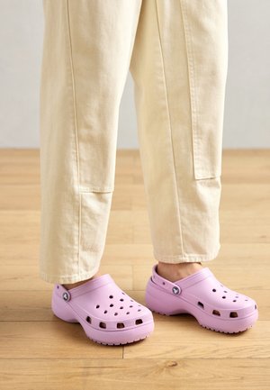 Sabots en caoutchouc violet avec des trous de ventilation, bout arrondi et bride arrière. Portés avec un pantalon beige clair sur un sol en bois.