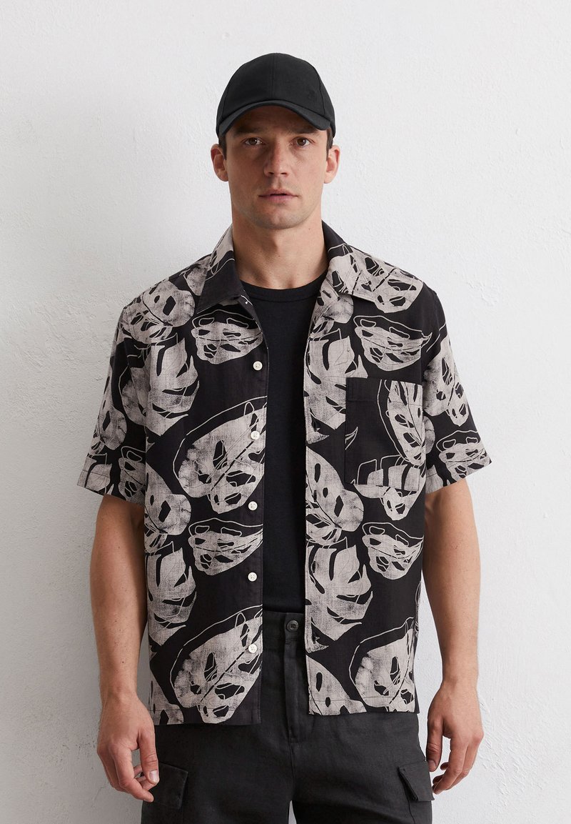 Hemd mit kurzen Ärmeln, das einen schwarzen Hintergrund mit großen weißen Blattmotiven zeigt. Es verfügt über eine Brusttasche und Knöpfe auf der Vorderseite.