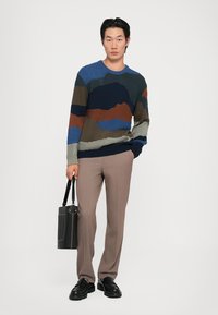 Pulover tricotat cu modele abstracte multicolore în nuanțe de navy, verde și ruginiu, combinat cu pantaloni bej și pantofi negri, ținând o geantă neagră.