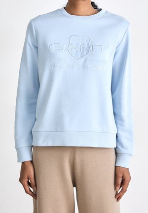 Personne portant un sweat-shirt GANT bleu clair avec logo embossé et un pantalon beige, debout devant un fond blanc uni.