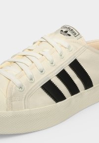 Κοντινή λήψη λευκού αθλητικού παπουτσιού Adidas με μαύρες ρίγες, λευκά κορδόνια και το λογότυπο της Adidas στη γλώσσα.
