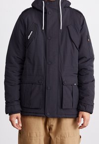 Schwarze gesteppte Jacke mit Kapuze, mit einer Reißverschlusstasche am Ärmel, Druckknöpfen und zwei Fronttaschen.