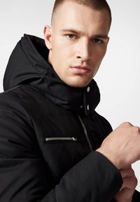 Czarna pikowana kurtka z wysokim kołnierzem, z metalowymi zamkami błyskawicznymi i zatrzaskami, widoczną teksturą przeszycia oraz przednią kieszenią.