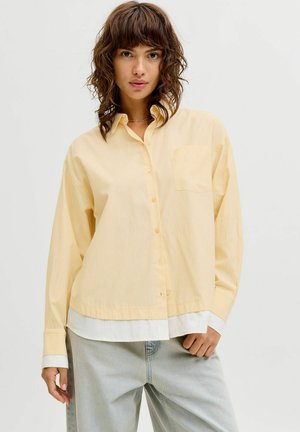 Nainen, jolla on kiharat ruskeat hiukset, pukeutunut löysään vaaleankeltaiseen nappipaitaan valkoisen topin ja vaaleansinisien farkkujen päällä yksinkertaista taustaa vasten.