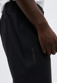 Gros plan sur la main d'une personne reposant à côté d'un pantalon noir de la marque Salomon avec une taille élastique.