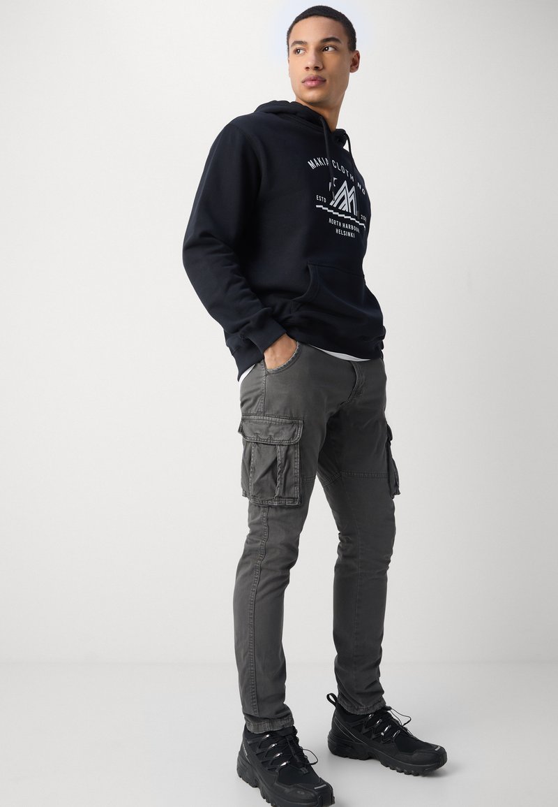 Sudadera con capucha negra con gráfico blanco, combinada con pantalones cargo gris oscuro con bolsillos laterales y zapatillas robustas negras.