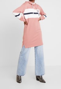 Robe à capuche rose avec des bandes blanches et navy sur la poitrine, dotée de fentes sur les côtés. Associée à un jean large bleu clair et des bottes à motifs.