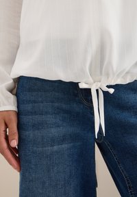 Blusa blanca con pliegues y dobladillo ajustable, combinada con unos vaqueros de denim azul oscuro. Primer plano de la blusa y los vaqueros, mostrando la textura de la tela.