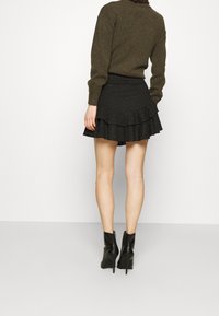 Jupe mini noire à volants avec une surface texturée ; associée à un pull vert foncé et des bottes noires à talons. Détails en tissu lisse visibles.