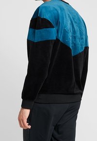 Svart och teal velour-tröja med en texturerad baksida, med svarta ränder över axlarna och en rundad halsringning.