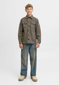 Camicia di flanella a quadrati marroni con due tasche sul petto, abbinata a jeans blu sbiaditi e scarpe beige, in piedi su uno sfondo semplice.