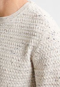 Gebreide trui in lichtbeige, met een textuurpatroon en subtiele vlekken. Klassieke ronde hals met ribbeldetails op de schouders.