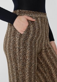 Leopardenmuster-Hosen mit glatter Textur und Seitentaschen. Sie zeigen ein braun-schwarzes Farbdesign auf einem hellbeigen Hintergrund.