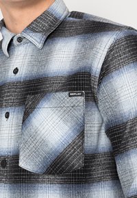 Primer plano de una camisa de cuadros azul, negra y blanca con botones negros y un bolsillo en el pecho etiquetado como "REPLAY."