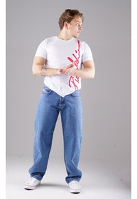 Weißes T-Shirt mit roter grafischer Schrift, kombiniert mit blauen Jeans. Das Modell trägt weiße Sneakers und zeigt einen lässigen, entspannten Look.