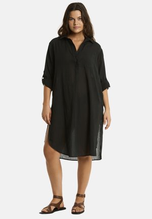 Ein Modell trägt ein lockeres schwarzes Hemdkleid mit hochgerollten Ärmeln und seitlichen Schlitzen, kombiniert mit braunen Sandalen mit Riemchen. Natürliches Haar, neutraler Hintergrund.