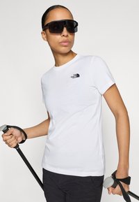 Λευκό κοντομάνικο μπλουζάκι με μαύρο λογότυπο στο στήθος, συνδυασμένο με μαύρο παντελόνι και κρατώντας μια ράβδο πεζοπορίας με γκρι λαβή.