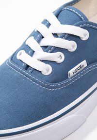 Κοντινή λήψη μπλε καμβά παπουτσιού Vans με λευκά κορδόνια, μεταλλικά ρούμπστικα, λευκή ραφή και λευκή λαστιχένια σόλα με ετικέτα λογότυπου.