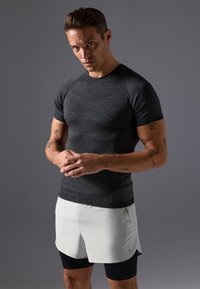 Schwarzes, kurzärmeliges Sportshirt aus strukturiertem Stoff, kombiniert mit weißen Shorts über schwarzen Kompressionsleggings. Das Modell posiert vor einem grauen Hintergrund.