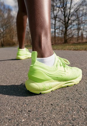 Osoba nosząca jasne, neonowozielone buty do biegania marki Under Armour oraz białe skarpetki, stojąca na utwardzonej ścieżce na zewnątrz, z drzewami w tle.