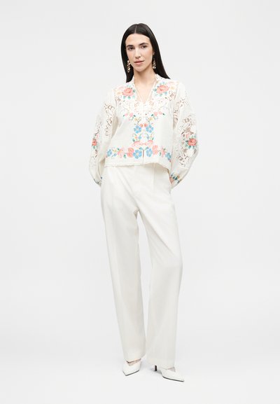 Γυναίκα που φοράει λευκή μπλούζα με πολύχρωμη κεντητή ανθοκομική διακόσμηση και δαντελωτά μανίκια, συνδυασμένη με λευκά παντελόνια φαρδιάς γραμμής και λευκές ψηλοτάκουνες γόβες.