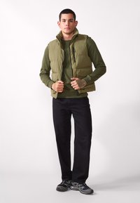 Olivenfarget polstret vest med stående krage, frontglidelås og to lommer, lagd over en matchende skjorte med lange ermer og kombinert med svarte jeans.