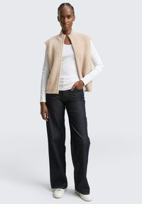 Beige gebreide vest met hoge kraag, gecombineerd met een witte blouse met lange mouwen en donkere wijde jeans. Lichte sneakers maken de outfit af.