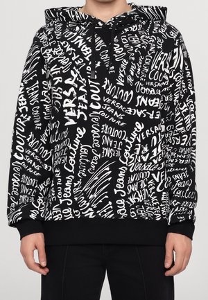 Zwarte hoodie met wit all-over tekstpatroon met verschillende lettertypen van "Versace Jeans Couture", gedragen door persoon zichtbaar van nek tot heupen.
