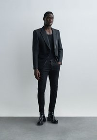Muž v čiernom na mieru ušitom saku, čiernej košeli, čiernych úzkych nohaviciach a lesklých čiernych topánkach stojaci pred svetlosivou stenou.